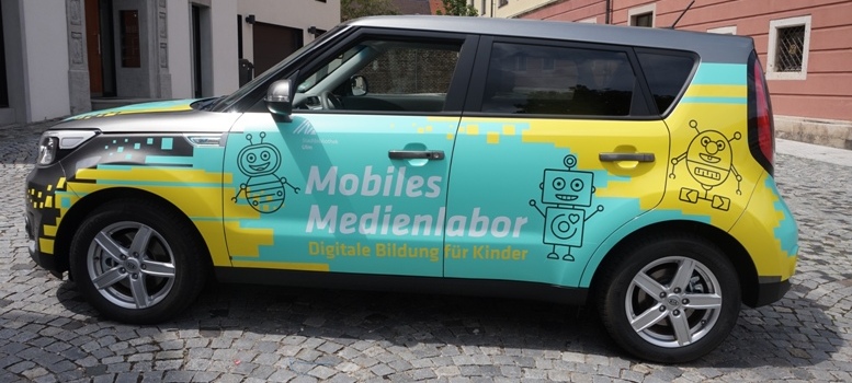 Ein türkis-gelb beklebtes Auto von der Seite mit der Aufschrift "Mobiles Medienlabor. Digitale Bildung für Kinder" und gezeichneten Robotern
