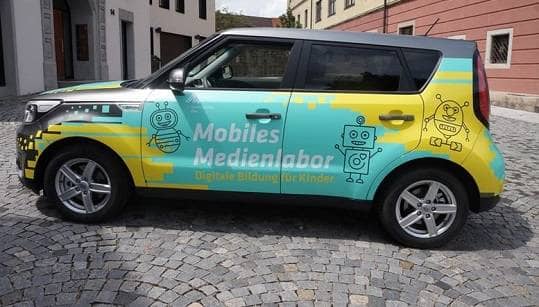 Ein türkis-gelb beklebtes Auto von der Seite mit der Aufschrift "Mobiles Medienlabor. Digitale Bildung für Kinder" und gezeichneten Robotern